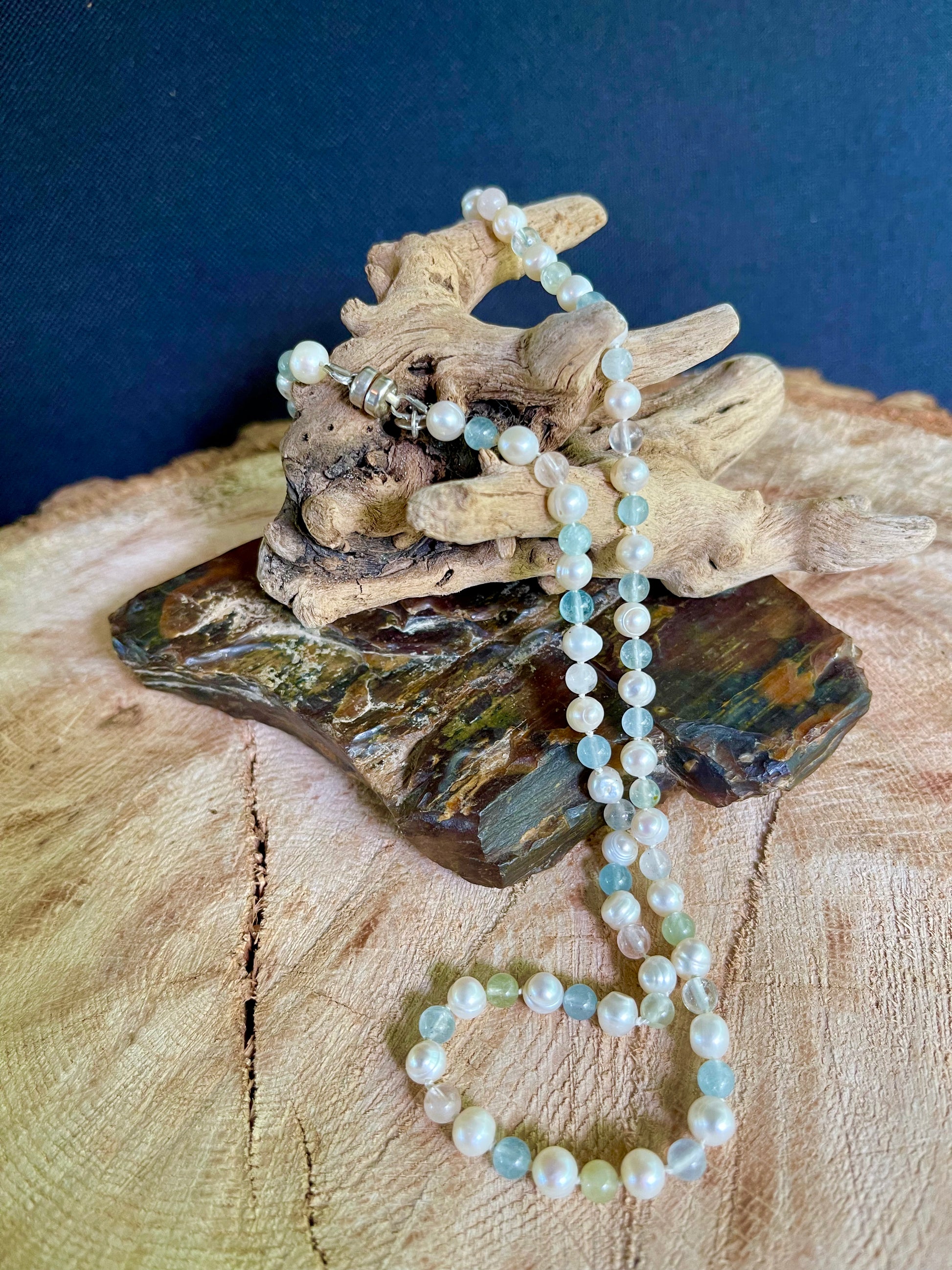 Pearl and Aventurine bead necklace draped over a piece of driftwood on a wooden surface with a dark background