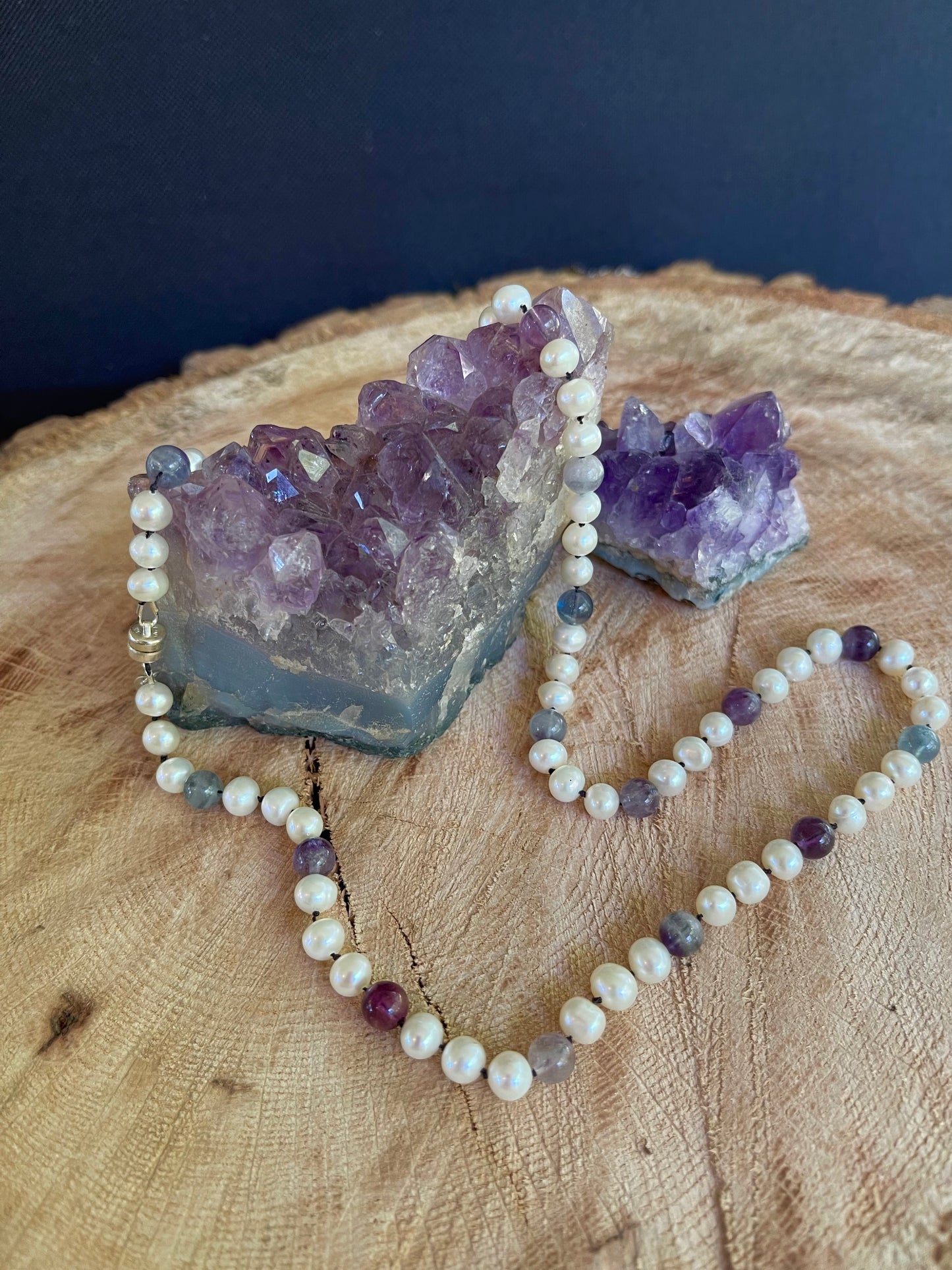White pearls and Fluorite beaded necklace on black Silk on a wooden surface with amethyst crystals.